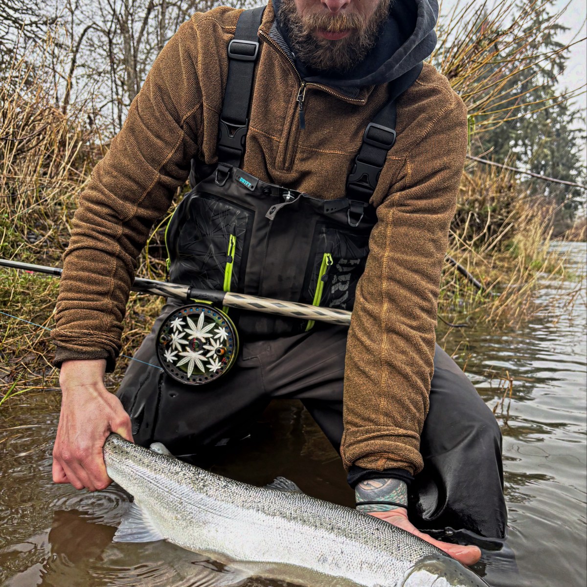 Man in waders holding a large fish by a riverbank.