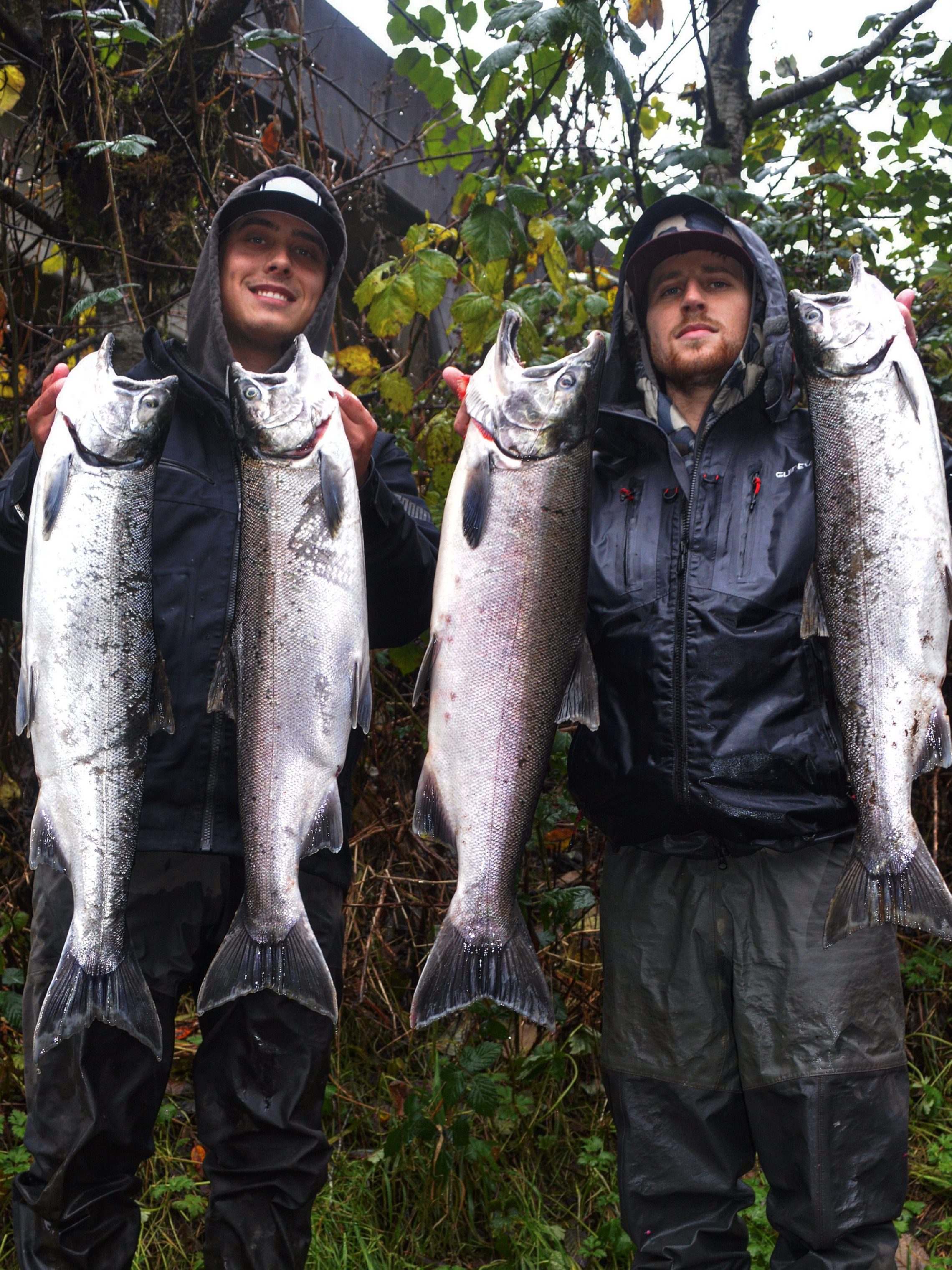 Two anglers proudly hold large fish in a forested area.