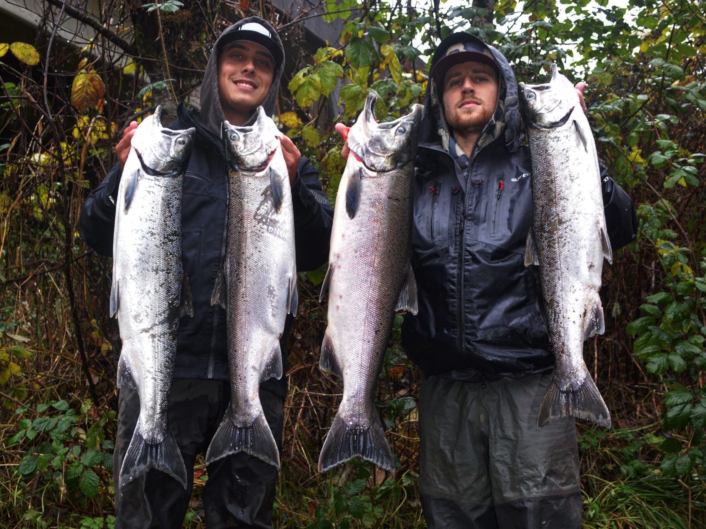 Two anglers proudly hold large fish in a forested area.