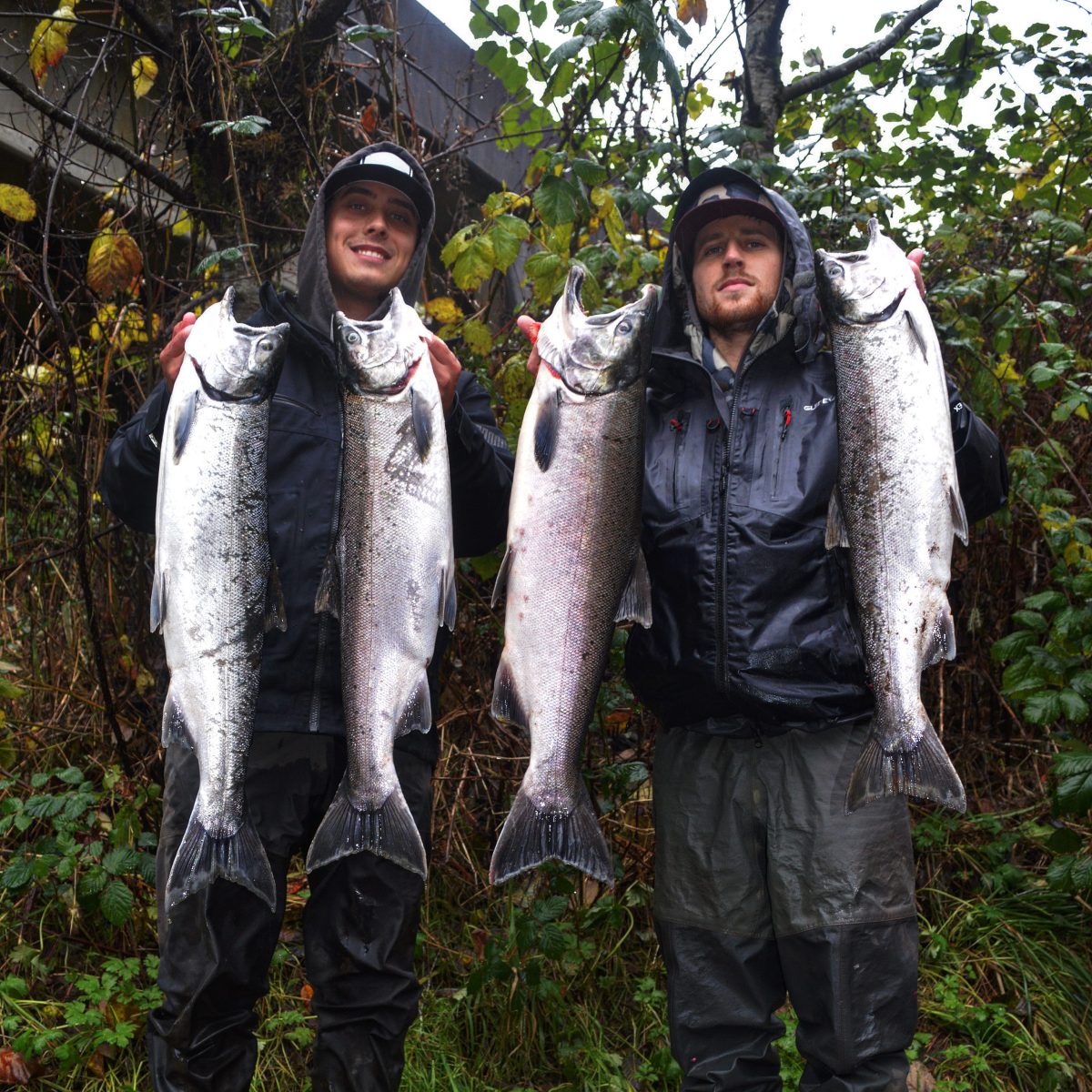 Two anglers proudly hold large fish in a forested area.