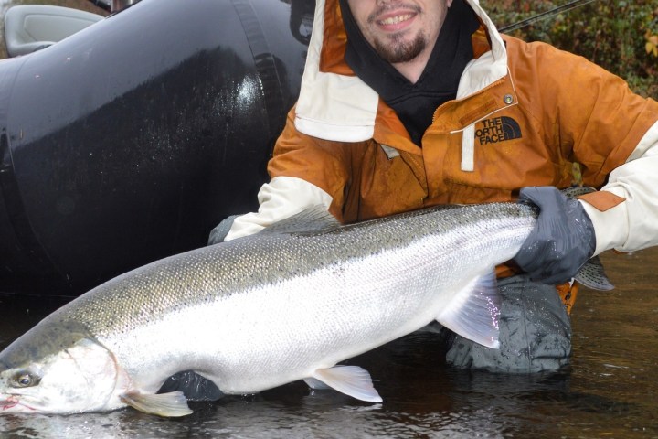 Person in brown jacket holding a large fish near water, with a black inflatable boat in the background.