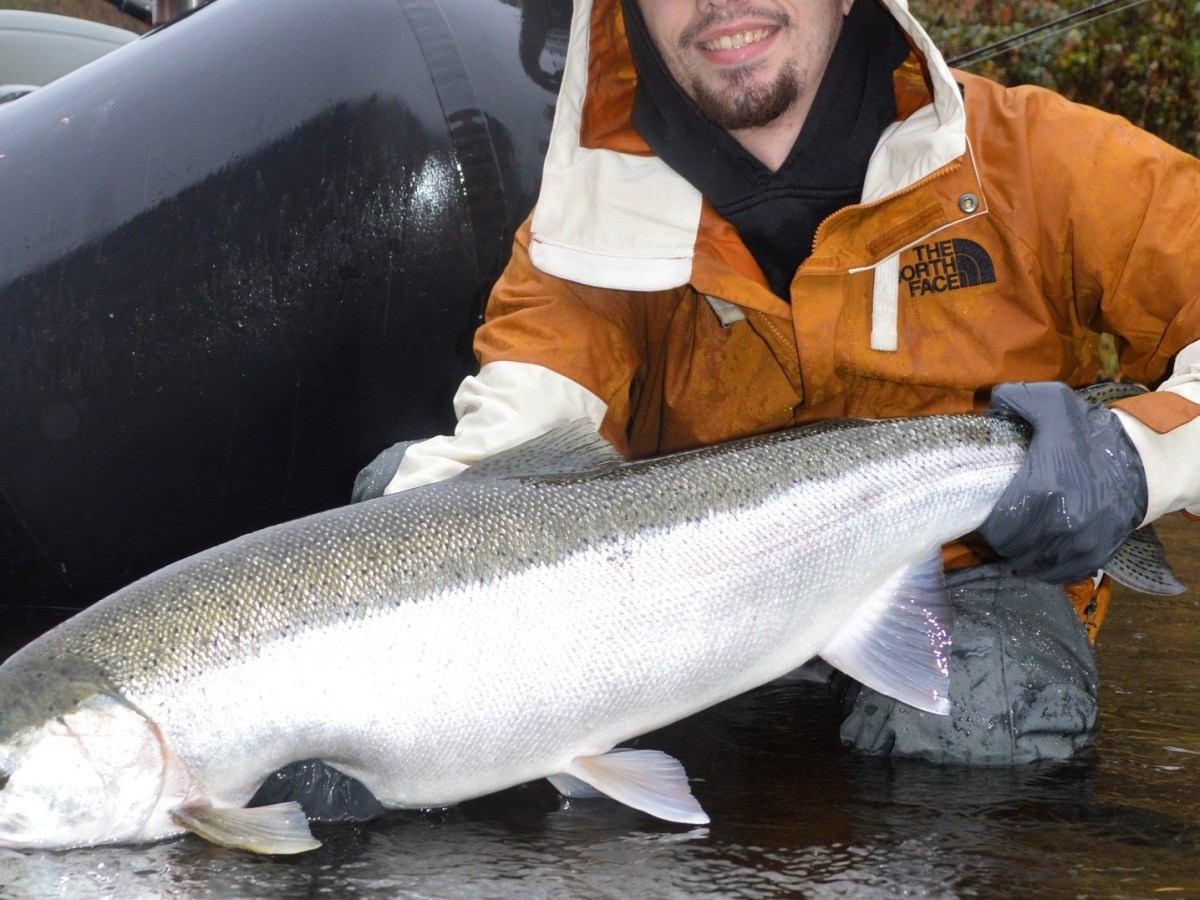 Person in brown jacket holding a large fish near water, with a black inflatable boat in the background.