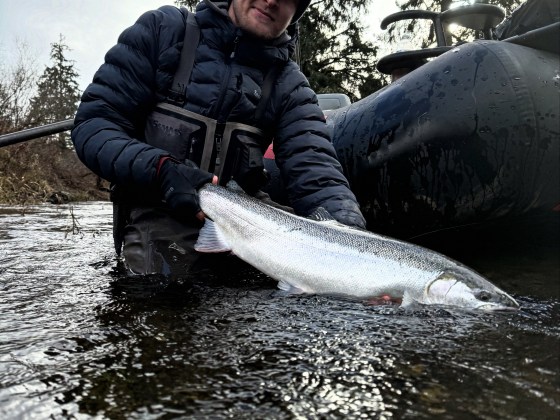Person in winter clothing holding a large fish near a boat on a river.