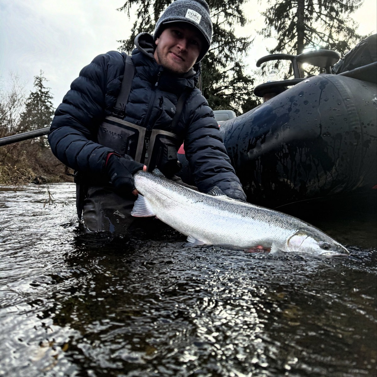 Person in winter clothing holding a large fish near a boat on a river.