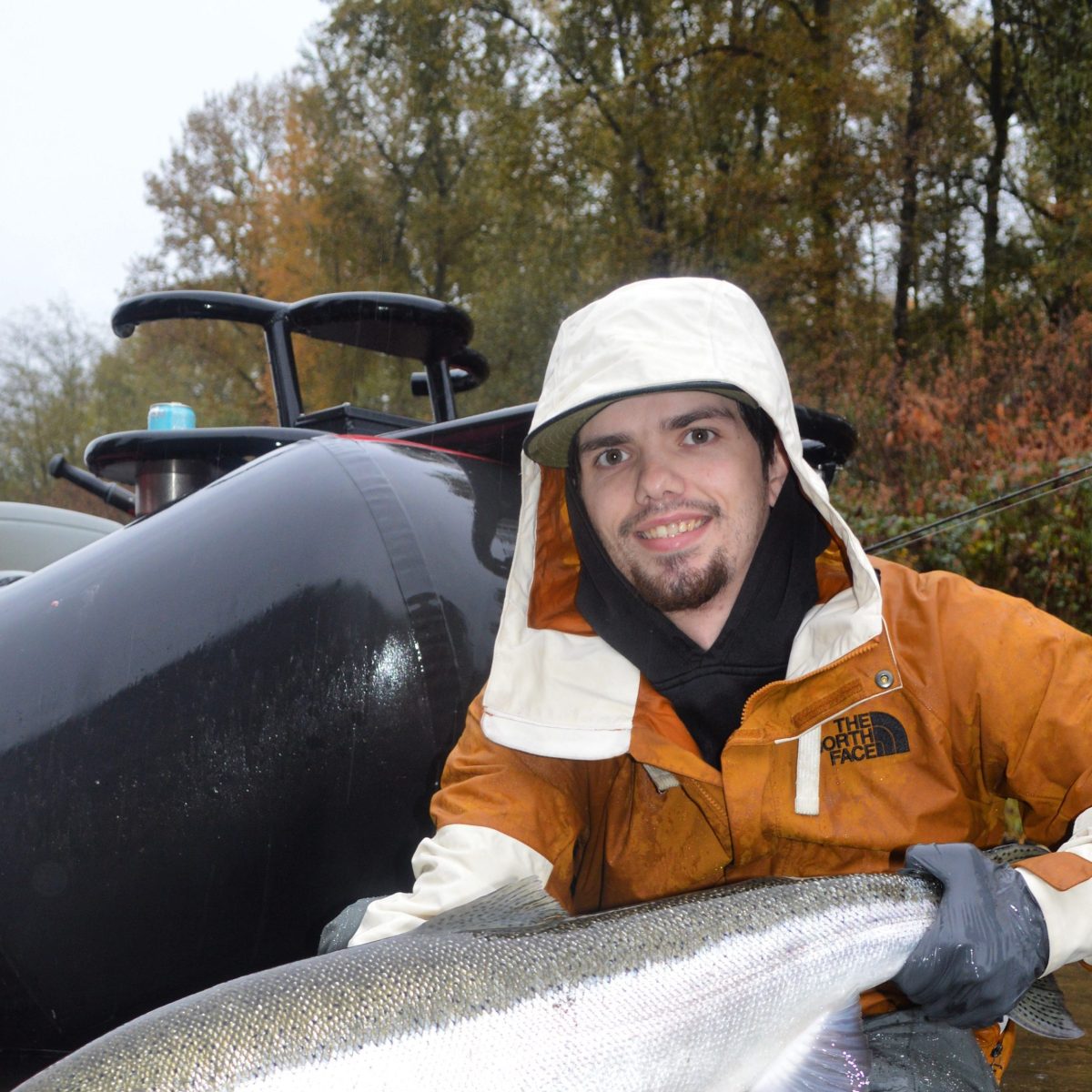 Person in hooded jacket holding a large fish by a river with a boat in the background.