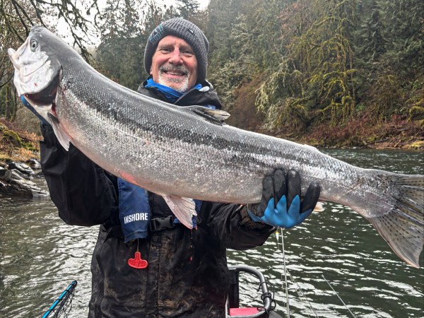 Person in winter clothing holds large fish on a boat in a forested river area.
