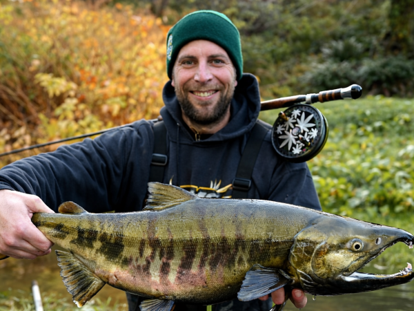 Man in outdoor gear holding a large fish near a river with autumn foliage.