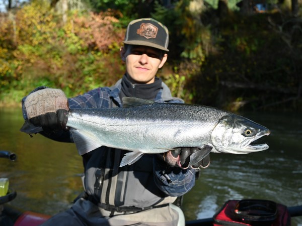 Person holding a large fish in both hands near a river, with autumn foliage in the background.