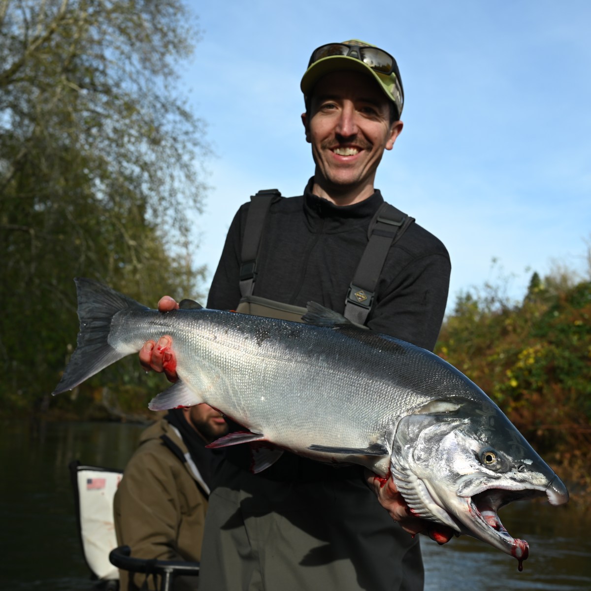 Man in outdoor gear holding a large fish with trees and water in the background.