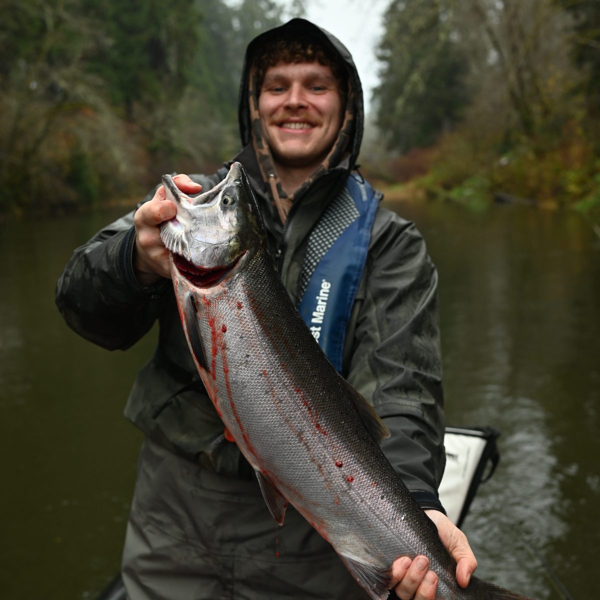 Man in rain gear holding large fish on a boat in a forested river.