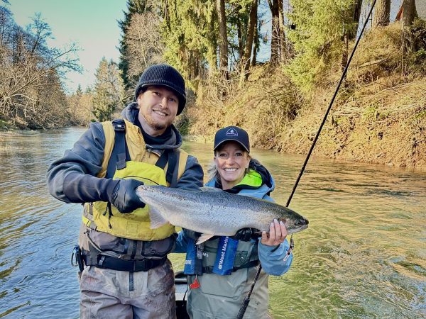 Two people on a boat holding a large fish, surrounded by a forested river scene.