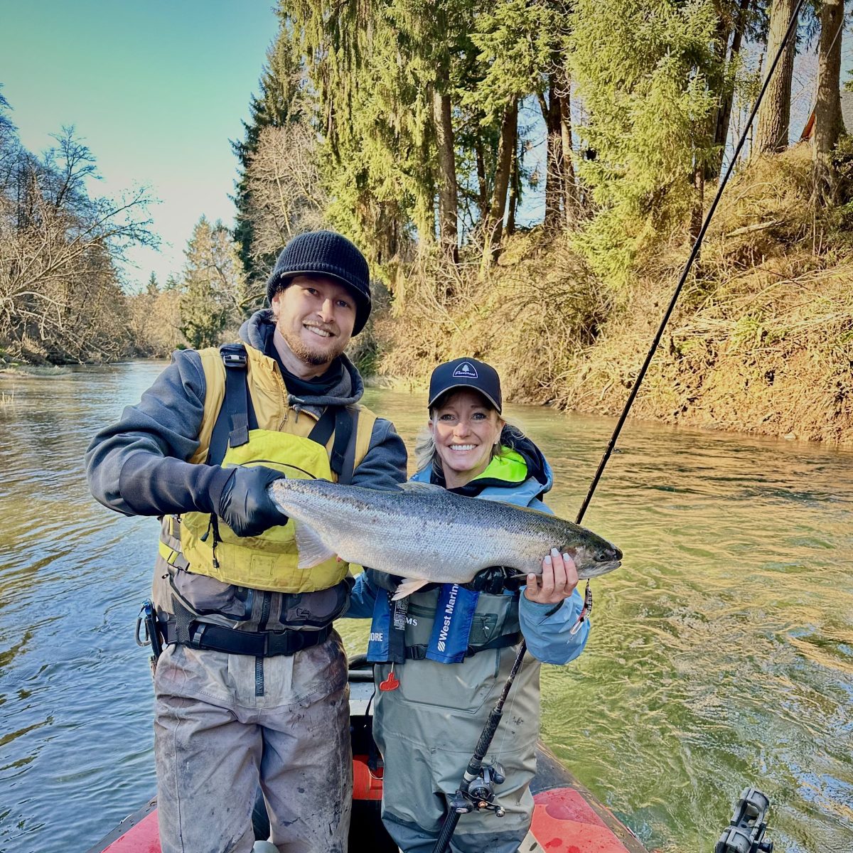 Two people on a boat holding a large fish, surrounded by a forested river scene.