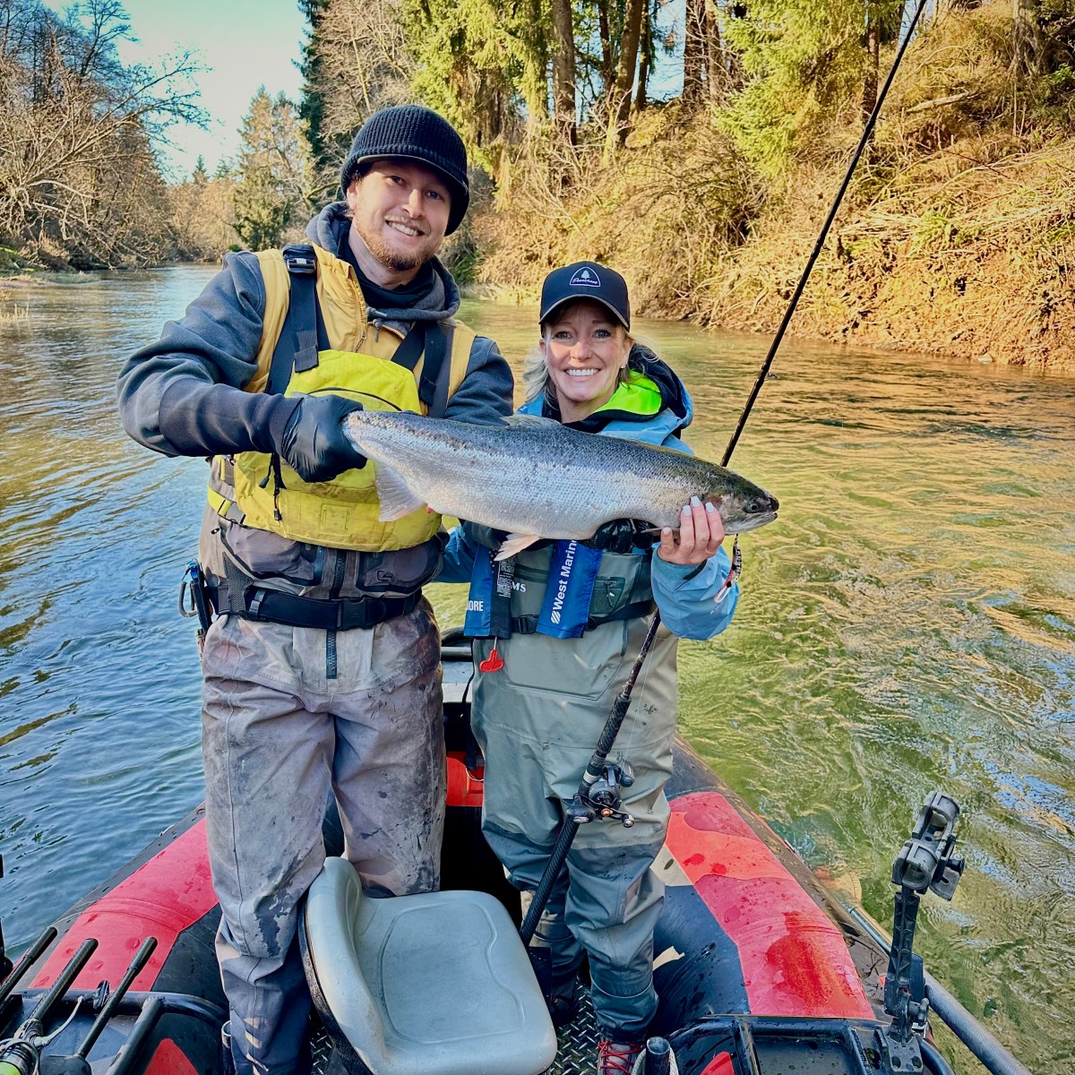 Two people on a boat holding a large fish, surrounded by a forested river scene.
