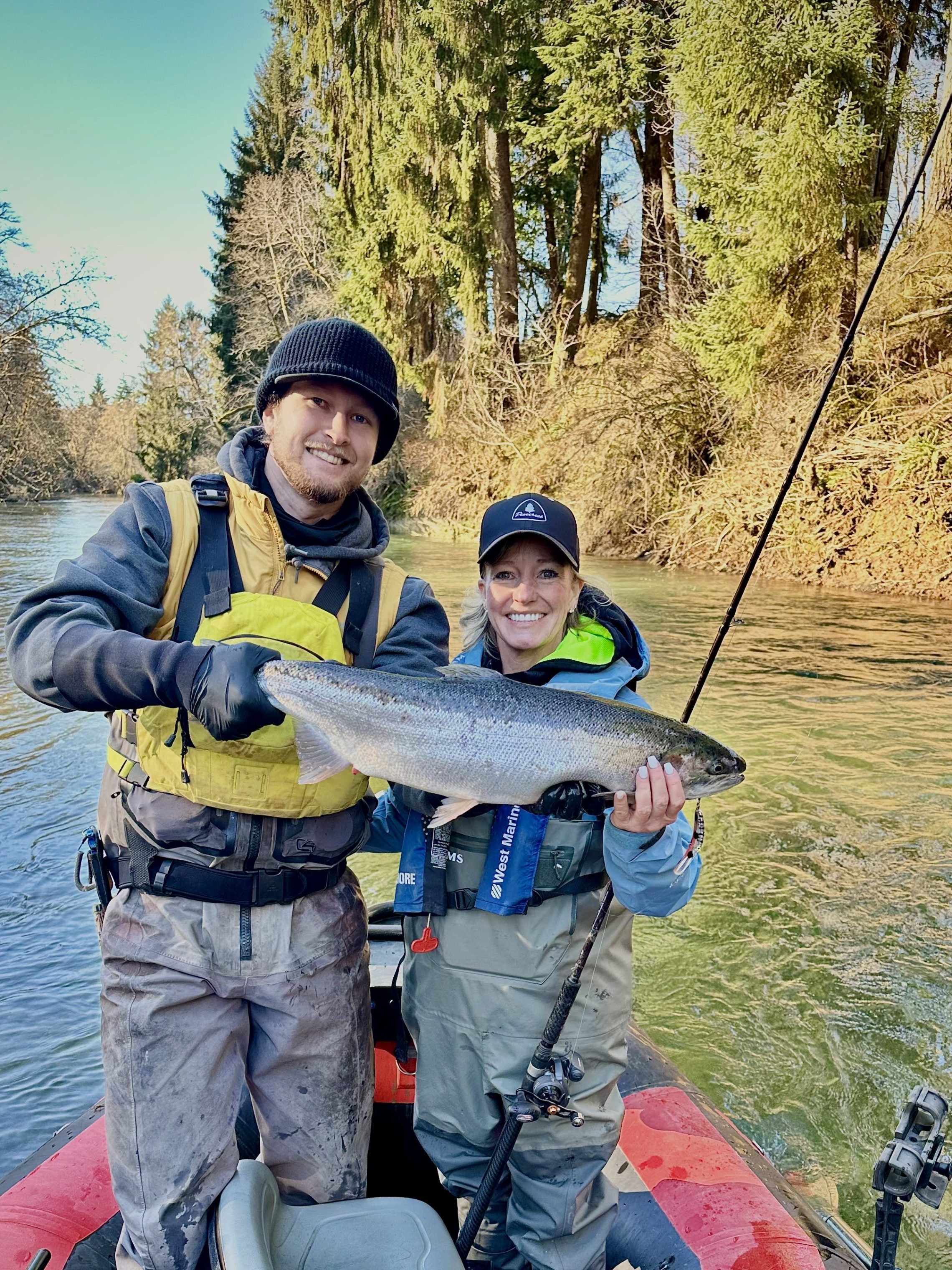 Two people on a boat holding a large fish, surrounded by trees.