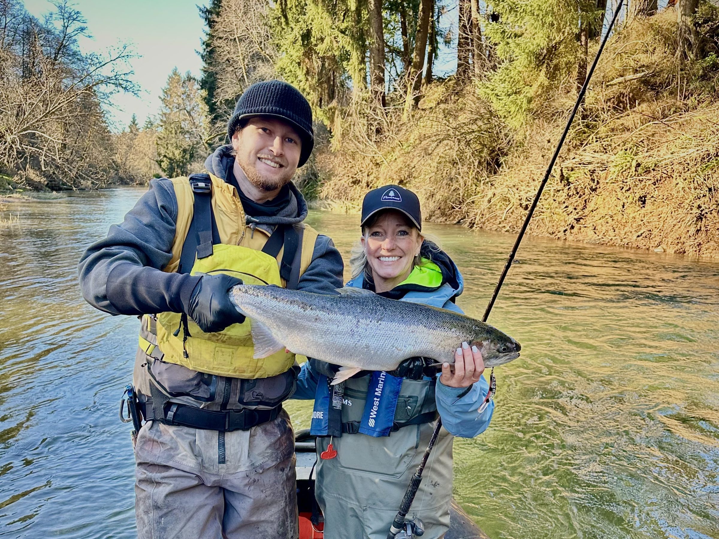 Two people on a boat holding a large fish, surrounded by trees.