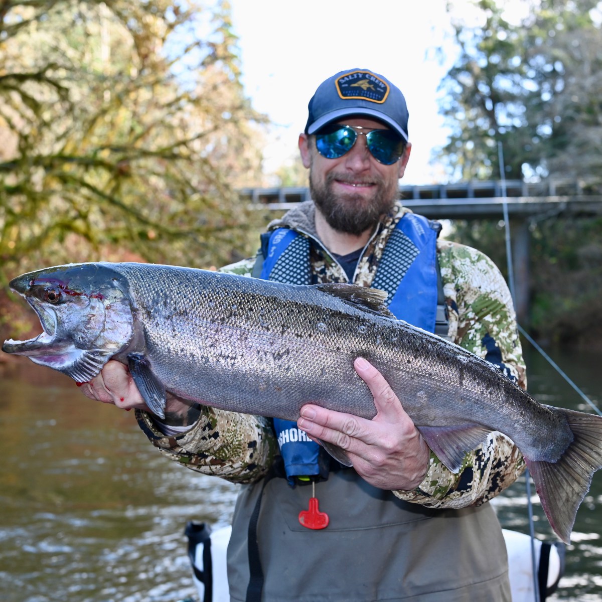 Bearded man holding a large fish by a river, wearing sunglasses and a hat.