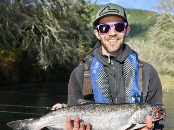 Person in a boat holding a large fish, wearing sunglasses and a cap, with trees in the background.