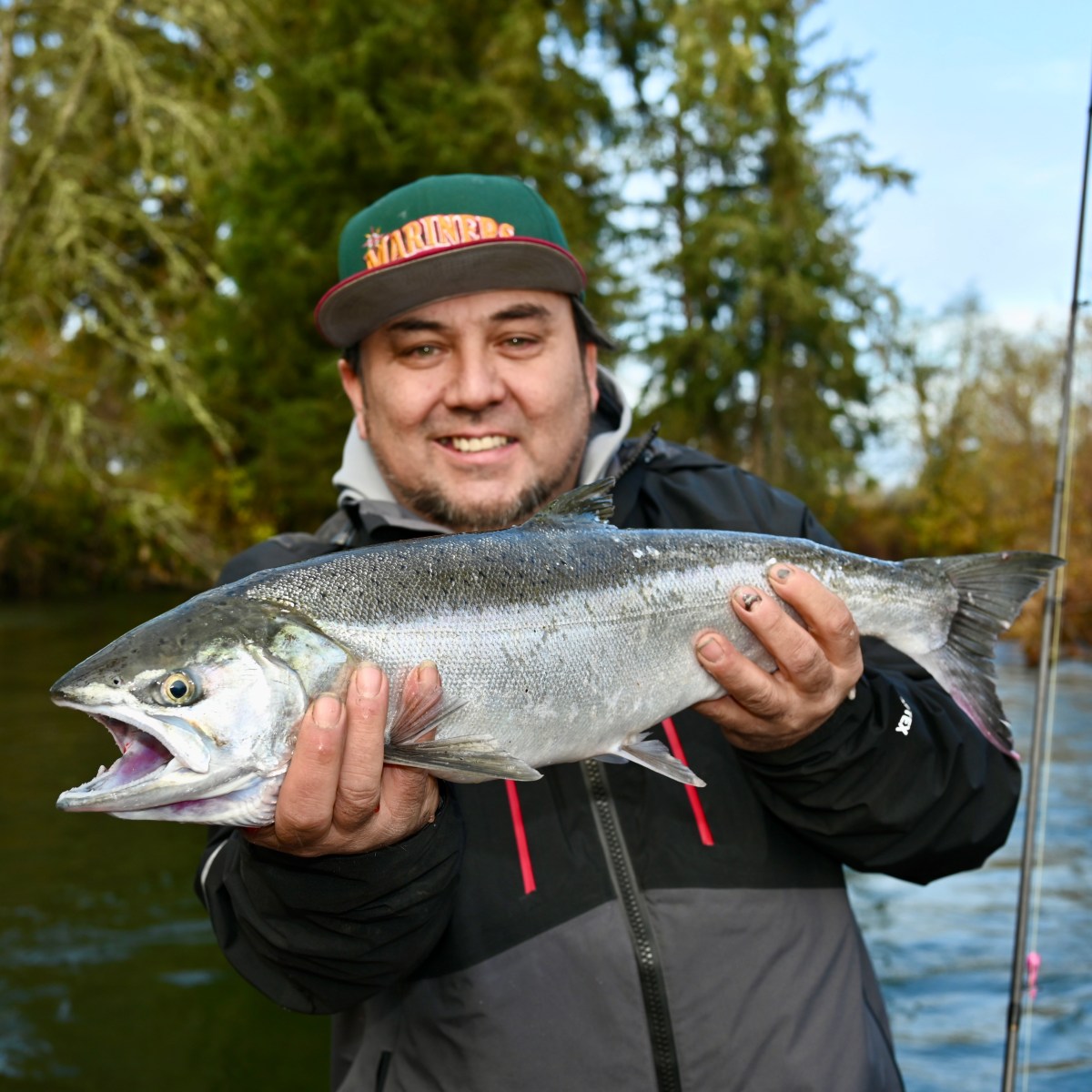 Person holding a large fish with open mouth by a river, wearing a cap and jacket, with trees in the background.