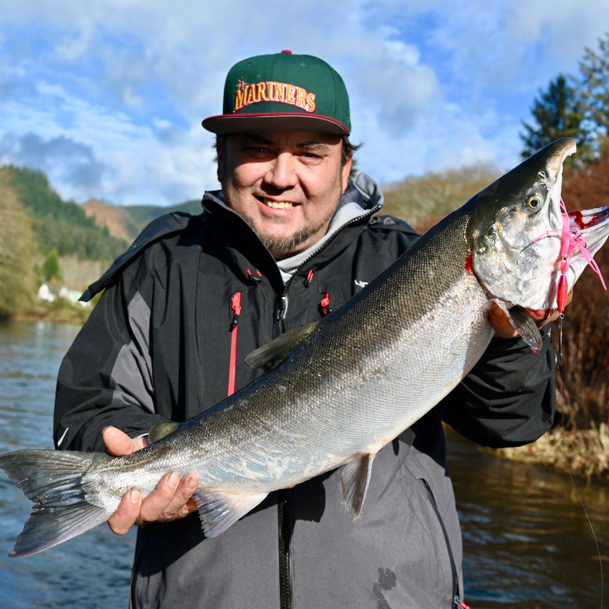 Person in a cap holding a large fish near a river with trees in the background.