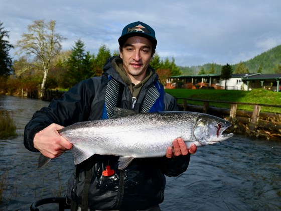 Person holding a large fish near a river with trees and a house in the background.