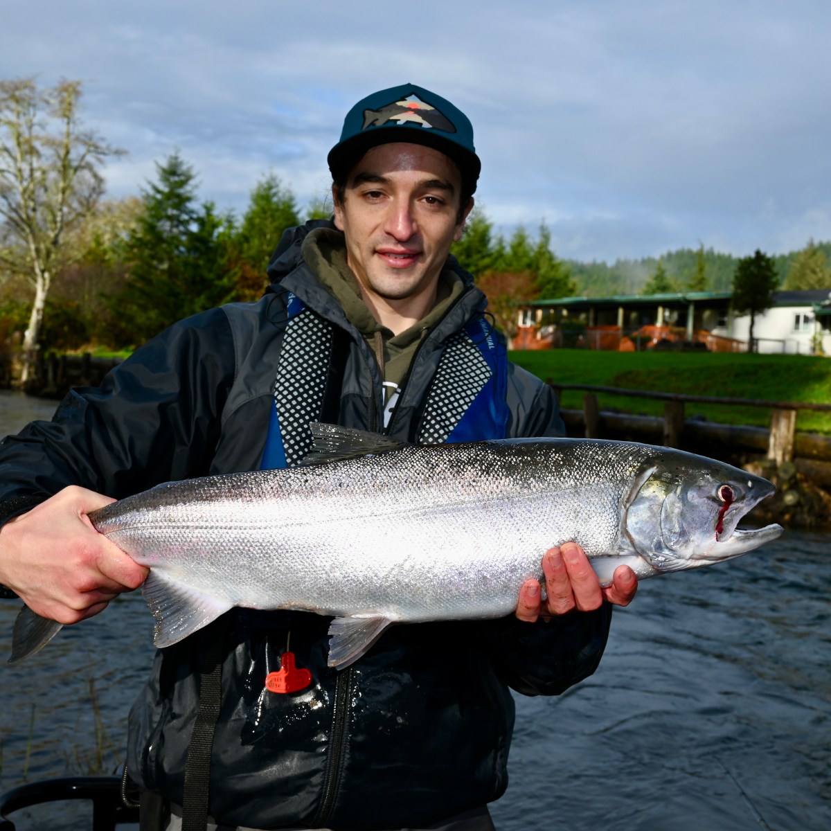 Person holding a large fish near a river with trees and a house in the background.
