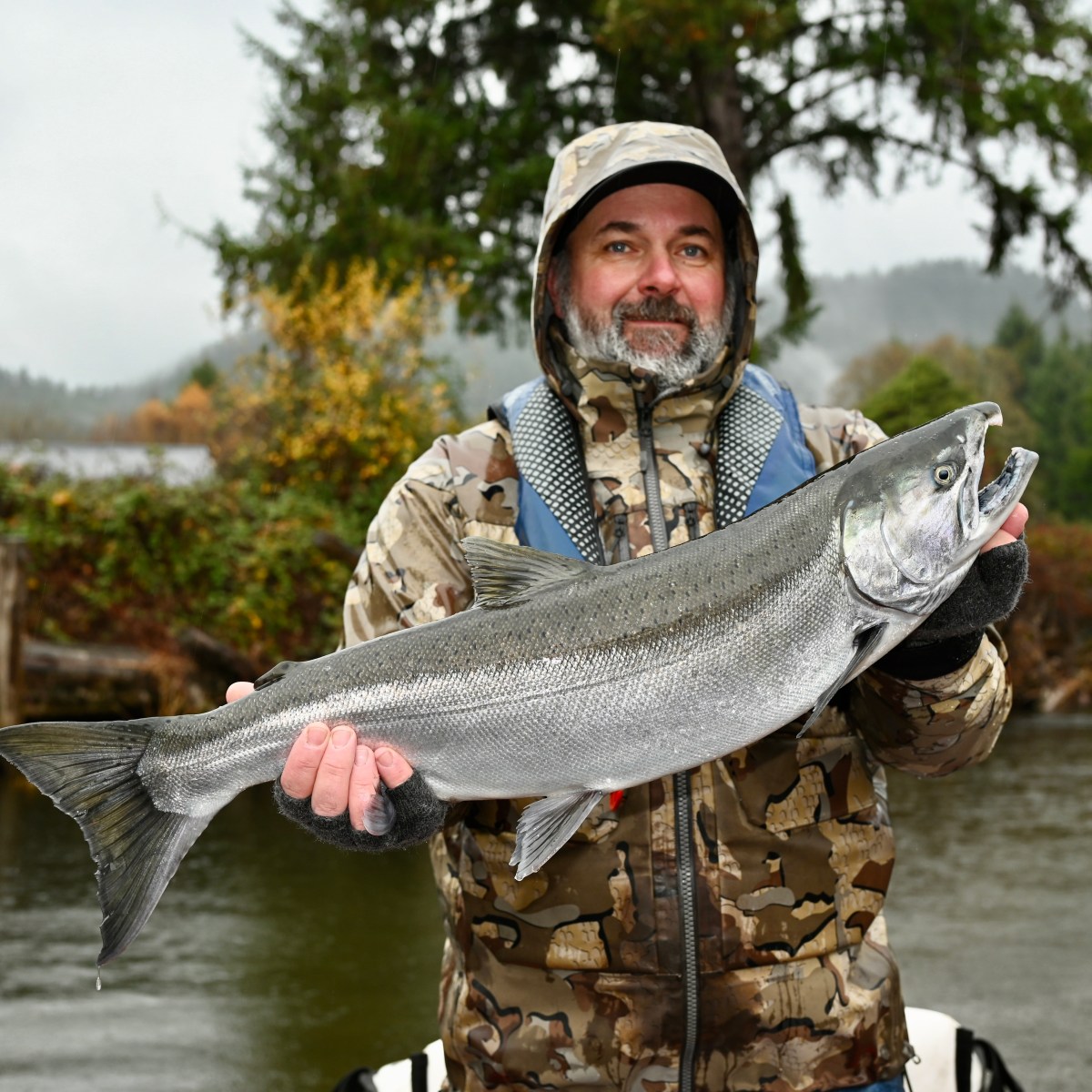 Person in camo jacket holds a large fish by the river with trees in the background.