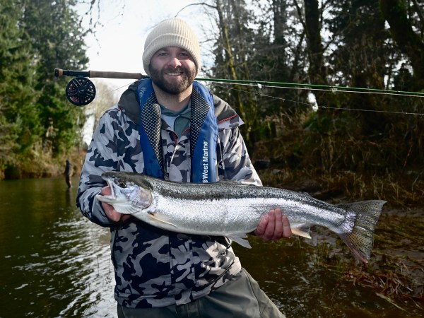 Person holding a large fish, wearing winter clothing and a fishing rod over the shoulder, standing by a river.