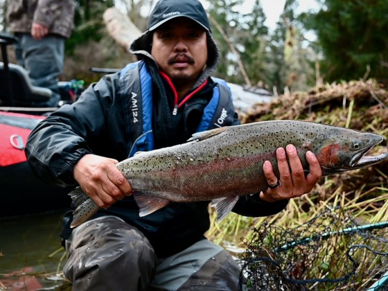 Person holding a large fish next to a river with a boat in the background.
