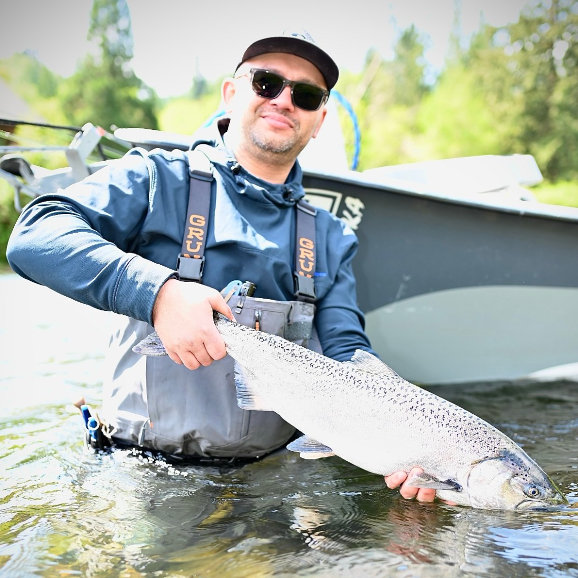 Person in fishing gear holds a large fish waist-deep in water near a boat.