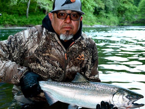 Person in camo jacket holds a large fish by a forest river.