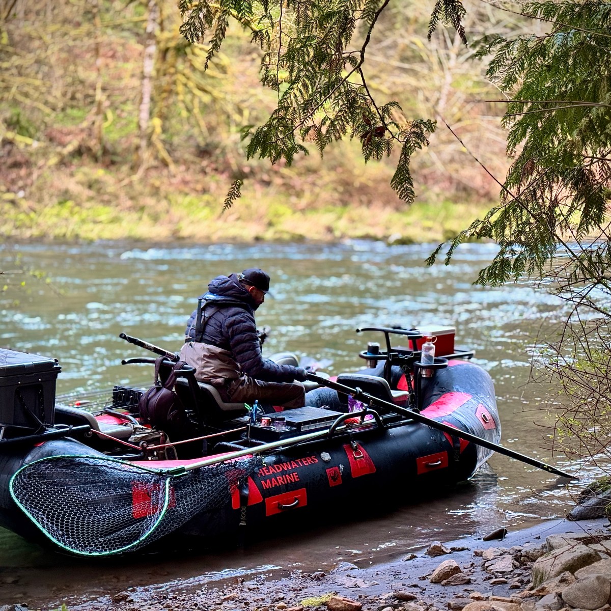 Person in winter clothing on an inflatable boat by a riverbank surrounded by trees.