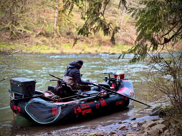 Person in a hooded jacket sitting on an inflatable boat by a riverbank, surrounded by trees.