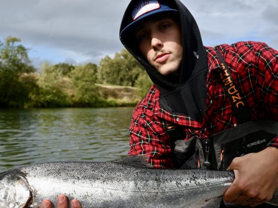 Person in red plaid shirt holding a large fish on a boat with a river background.
