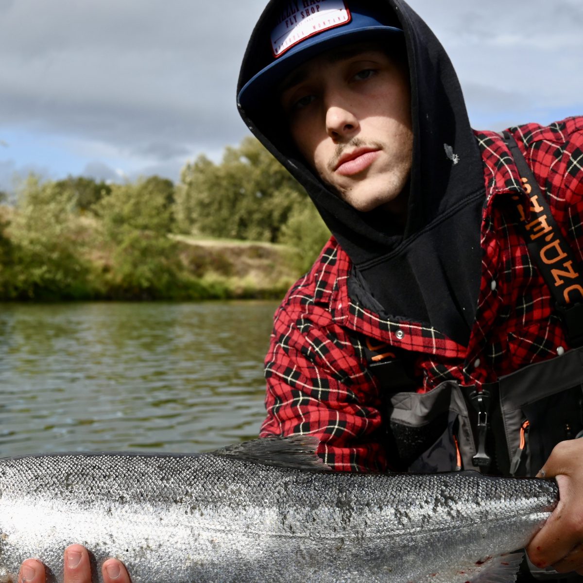 Person in red plaid shirt holding a large fish on a boat with a river background.