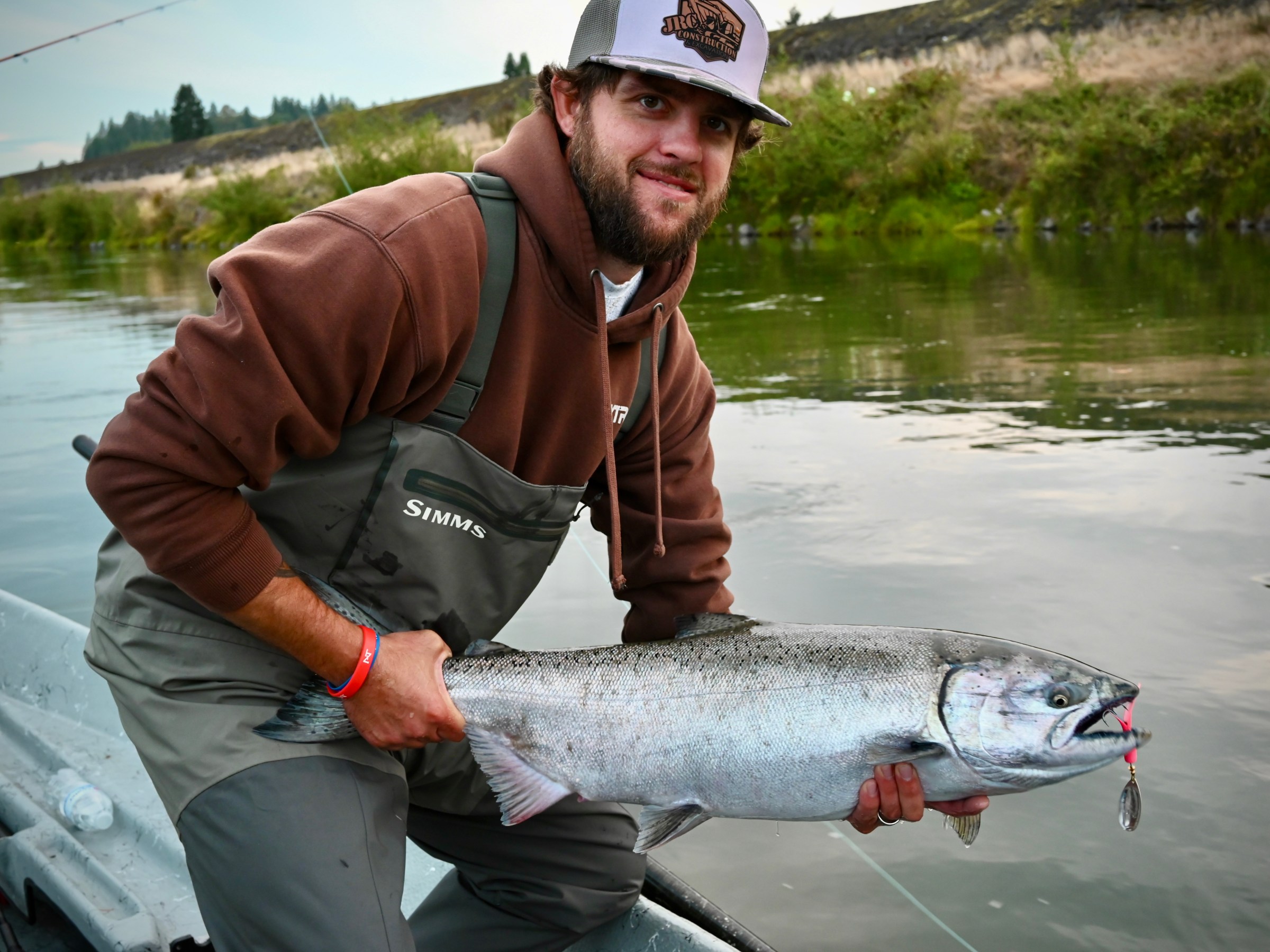 Person in a boat holding a large fish by a river, wearing a brown hoodie and gray cap.