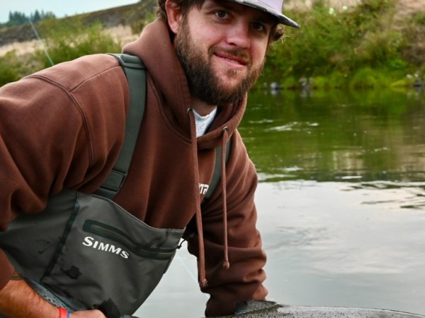 Man in brown hoodie and waders holding a fish by a river.