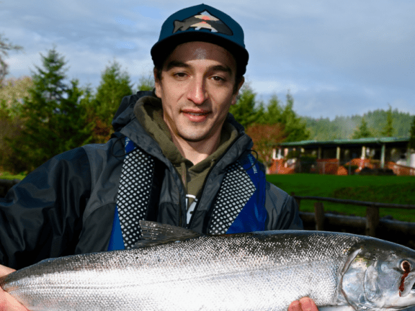 Person holding a large fish by a river with trees and buildings in the background.