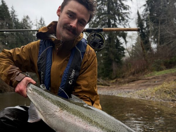 Person holding large fish in shallow river, wearing brown jacket and blue life vest, surrounded by trees.