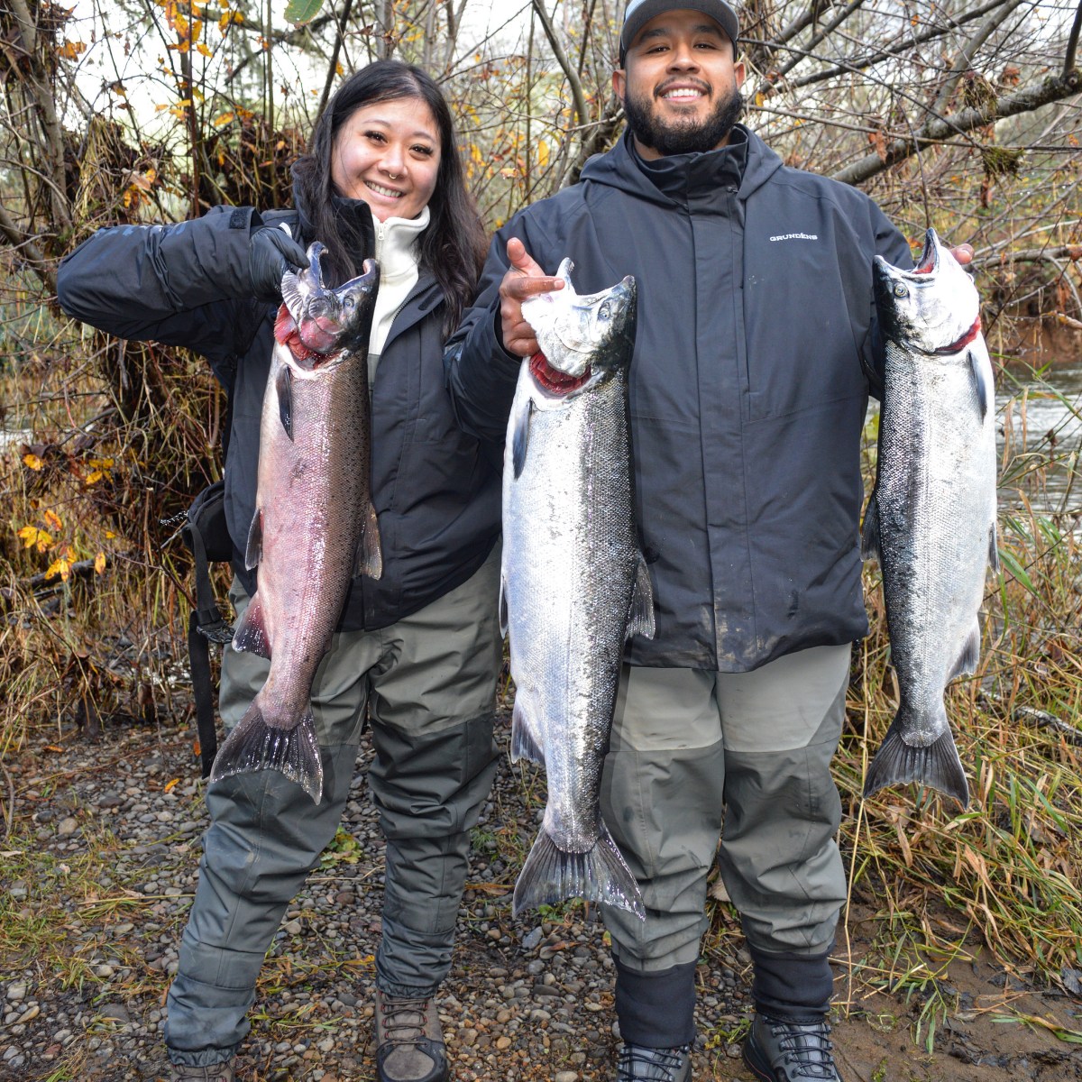 Two people in outdoor gear holding large fish in a wooded area.
