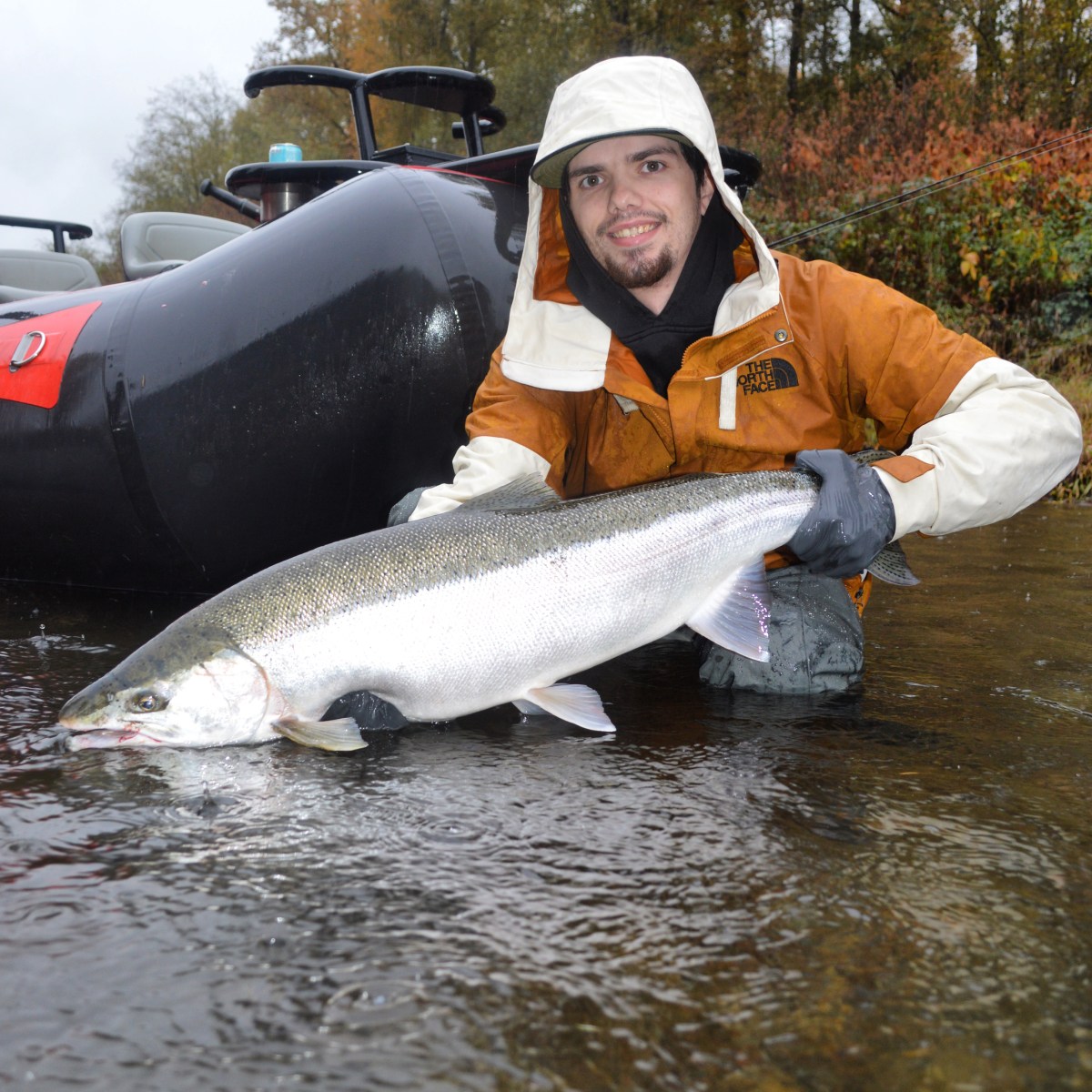 Person in raincoat holding large fish near a river with boat in background.