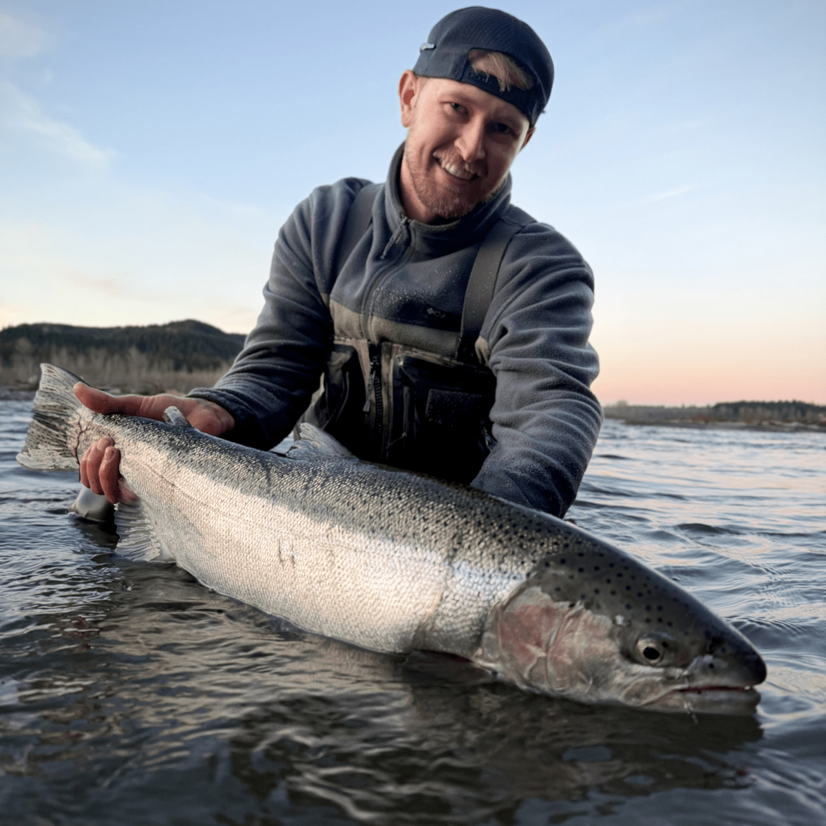 Person holding a large fish in shallow water, wearing a cap and jacket.
