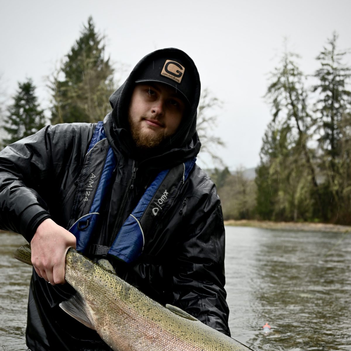Person in a black jacket and cap holding a large fish over water, with trees in the background.