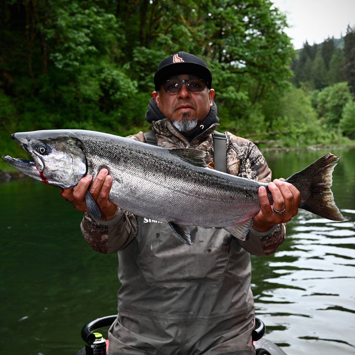 Man in camo gear holding a large fish on a boat with a forested background.