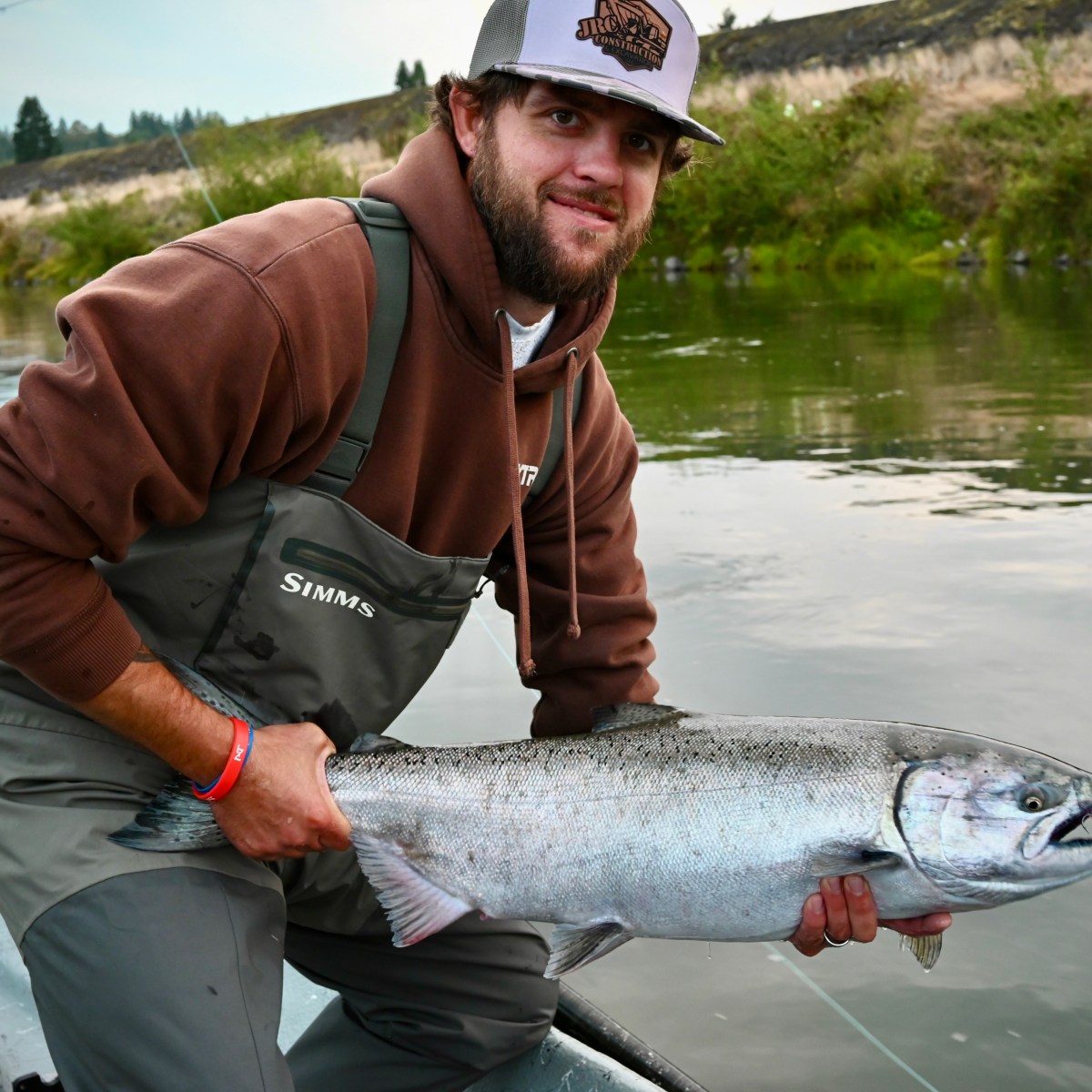 Person in brown hoodie and waders holding a large fish on a boat in a river.