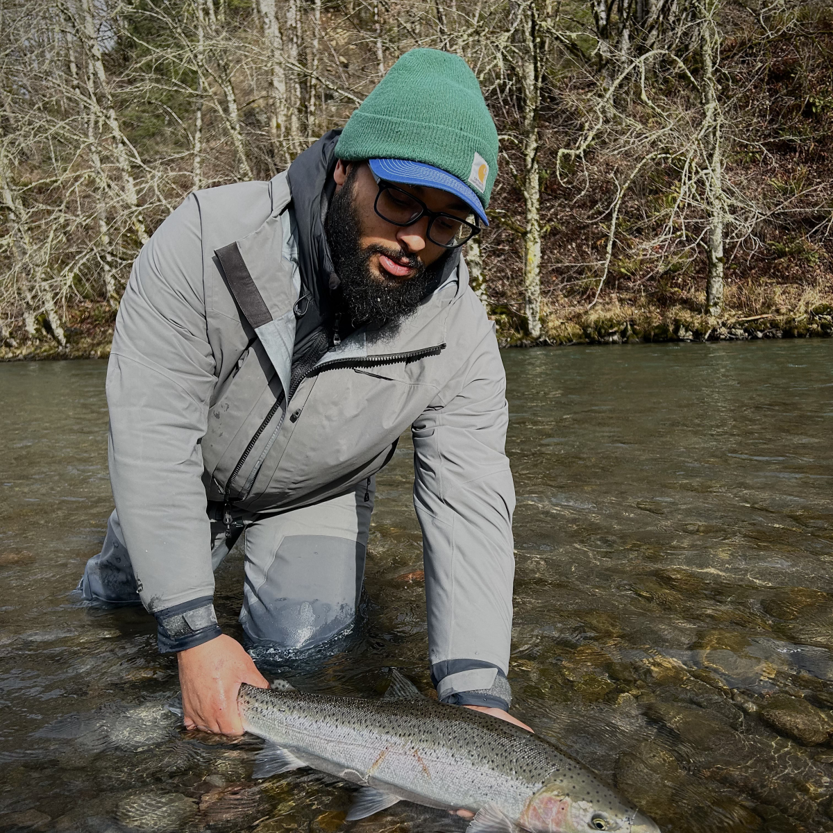Person in gray coat and green hat holding a fish in a river with trees in the background.