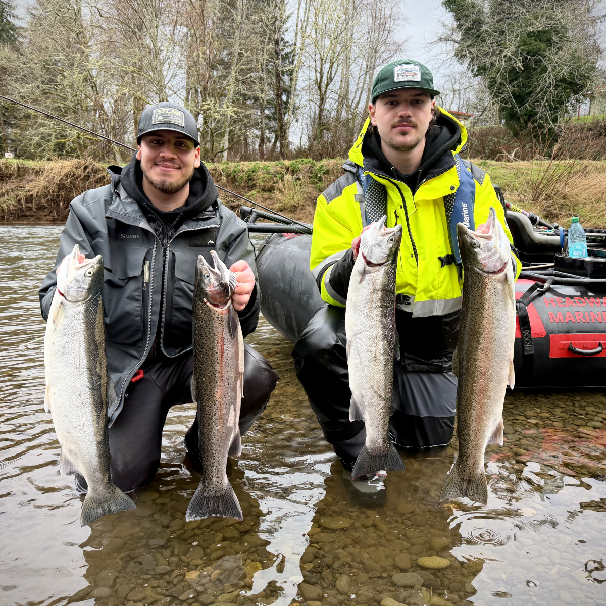 Two men kneeling in a river holding up fish, standing beside an inflatable boat.