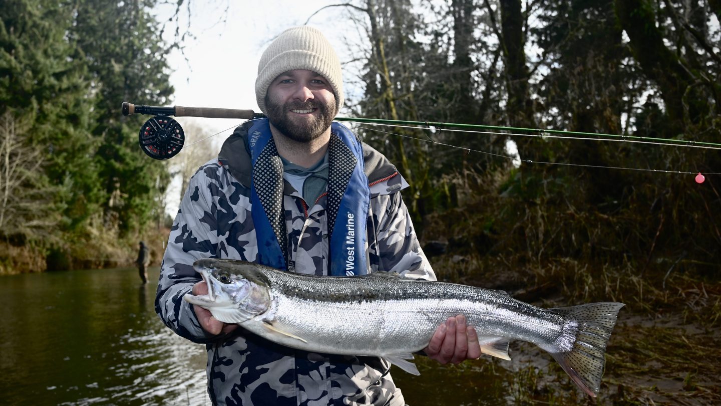 Man in camo jacket and beanie holding a large fish by a river, with a fishing rod on his shoulder.
