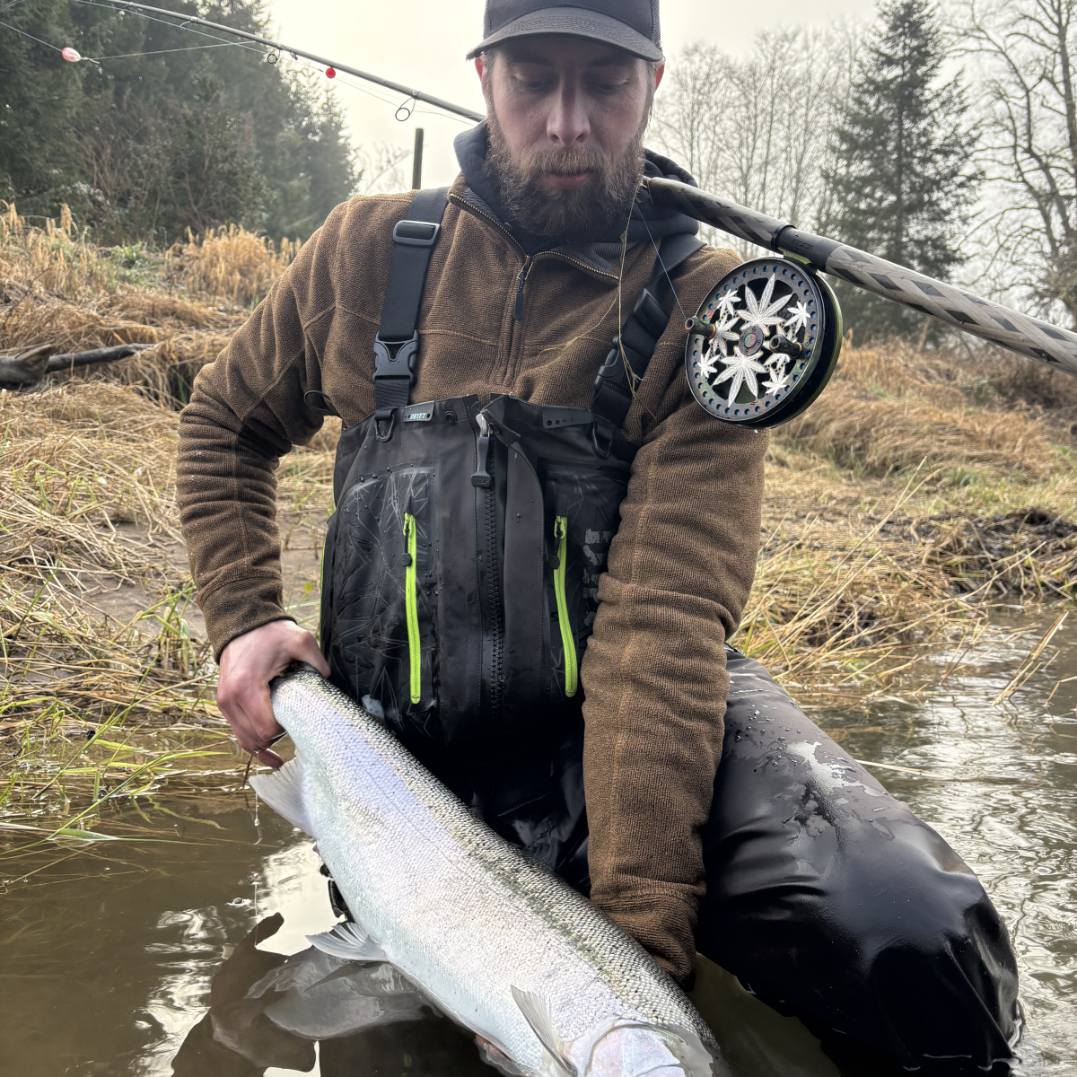 Man in waders holding a large fish by a river, with a fishing rod in the background.